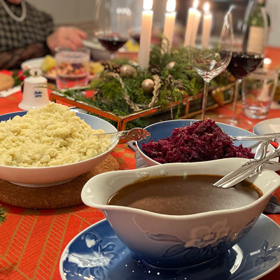 Heiligabend mit traditionellen Festgerichten und einem Tisch, gedeckt mit dem Bing & Gröndahl Weihnachtsrosen-Service. Servierschalen mit Rotkohl, Kartoffeln und Soße stehen auf einer roten Tischdecke, umgeben von Weihnachtsdekoration und Kerzenlicht.