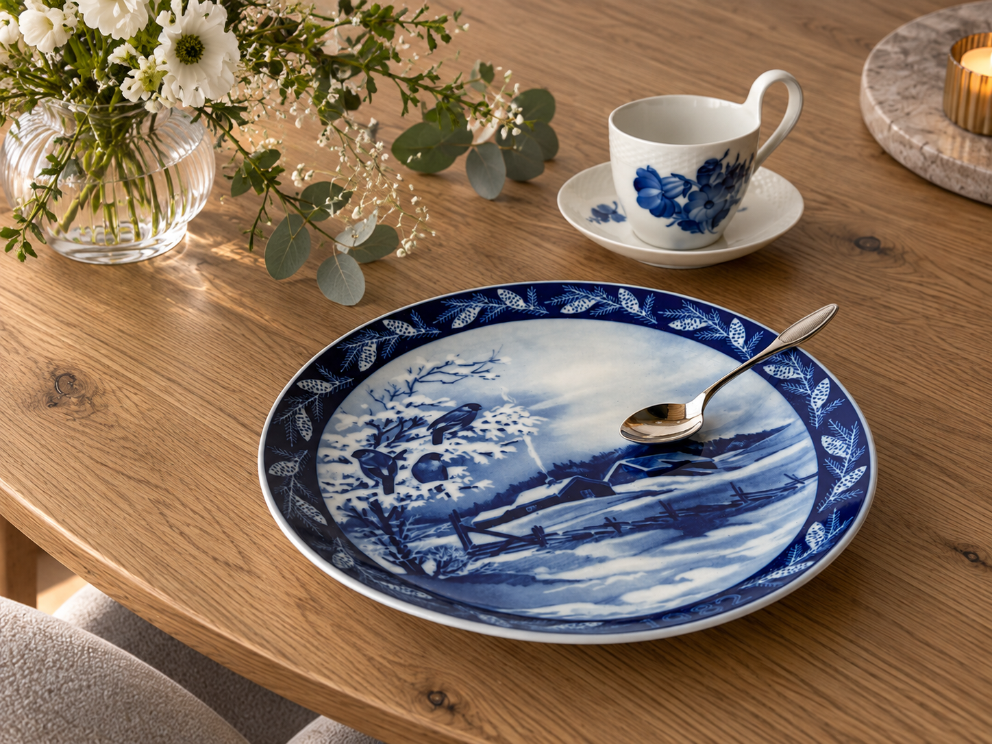 Heinrich Weihnachtsteller 1982 mit blau-weißem Wintermotiv, gedeckt auf Holztisch mit Blaublumen-Tasse, Löffel und Blumen.