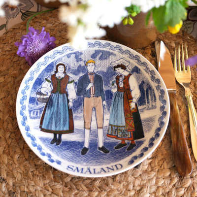 Ravn Teller mit einem Motiv von Volkstrachten aus Småland in Schweden, verwendet als Essteller in einer Tischdekoration mit geflochtener Tischunterlage, Blumen und goldenem Besteck.