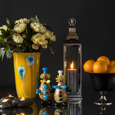 Zwei Clownfiguren aus Glas vor einer gelben Glasvase, Laterne und Obstschale