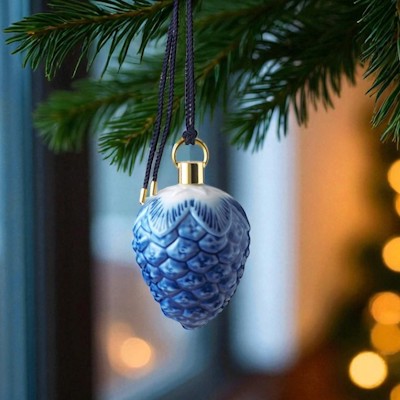 Royal Copenhagen Weihnachtsanhänger mit blauem Zapfenmotiv hängt an einem grünen Tannenzweig mit warmen Weihnachtslichtern im Hintergrund.