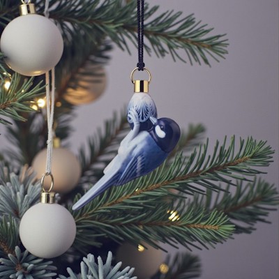 Royal Copenhagen Ornament 2021 mit blauem Meise hängt am Weihnachtsbaum zwischen grünen Zweigen, hellen Weihnachtskugeln und warmen Weihnachtslichtern.