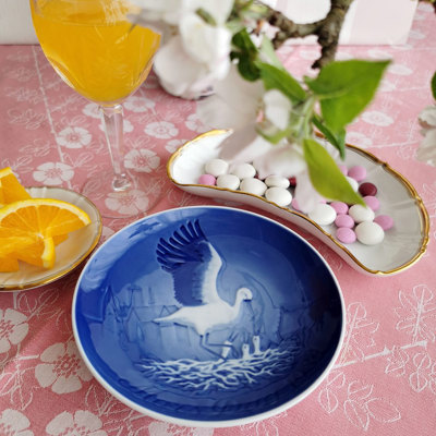 Bing & Gröndahl Muttertagsteller mit Storchenmotiv, verwendet in einer Muttertagstischdekoration. Auf rosa Tischdecke mit Blumen, Offenbach Geschirr, frischen Orangen und pastellfarbenen Schokoladenknöpfen.