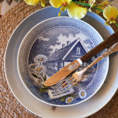 Tischdekoration mit einem Ravn Landschaftsteller, verwendet als Vorspeisenteller auf einem blauen Service. Eine geflochtene Tischunterlage und Blumen in Gelbtönen vervollständigen den persönlichen Look.