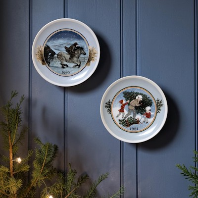 Zwei Tettau Weihnachtsteller mit Wintermotiven hängen an einer dunkelblauen Wand über Tannengrün und warmem Weihnachtslicht.