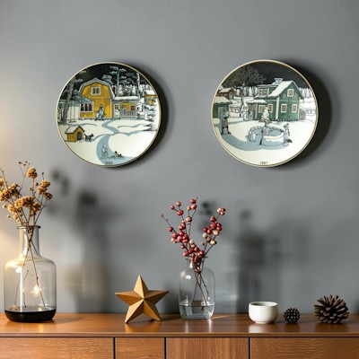 Zwei Arabia Weihnachtsteller mit farbenfrohen Wintermotiven hängen an einer grauen Wand über einem Sideboard mit Zweigen, Zapfen und Weihnachtsschmuck.